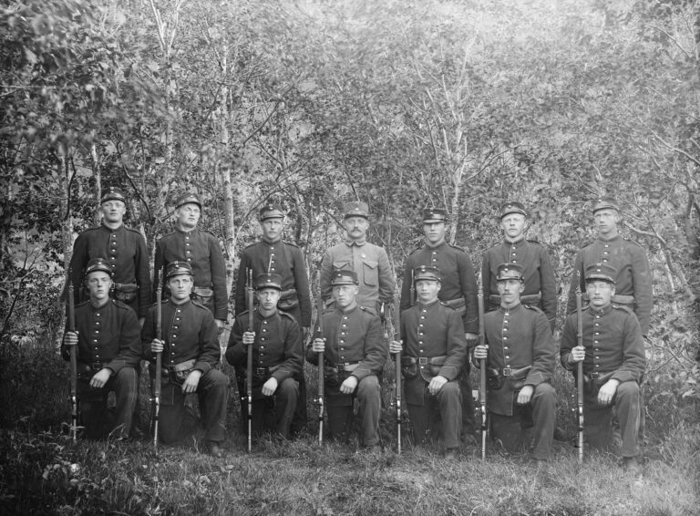 Historic Militia Training Unit Portrait | Early Black-and-white historic portrait of an early 20th century militia or military training unit posing outdoors with rifles in uniform formation among trees.