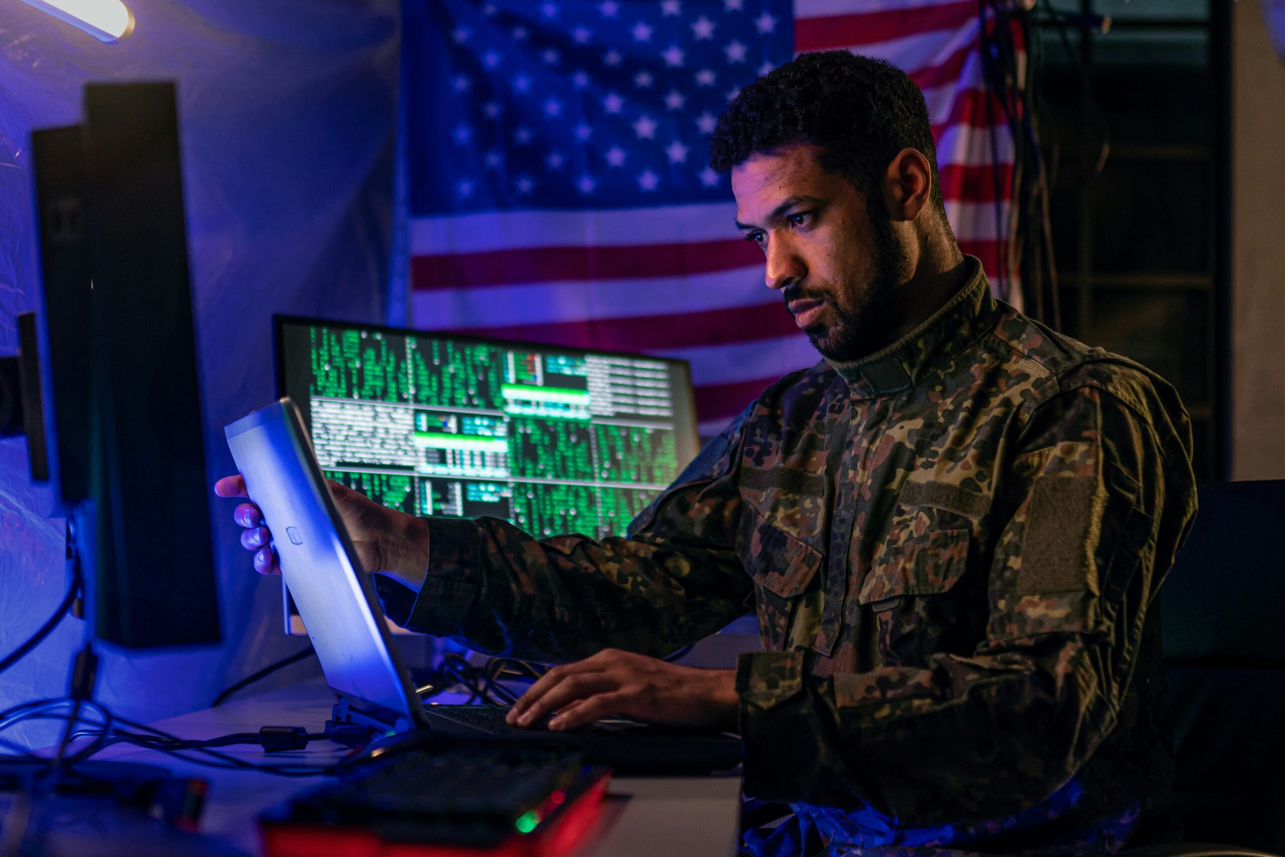 Member of the 55th Light Foot Battalion monitoring communications and digital systems during emergency preparedness and field operations training with American flag backdrop. SEO Caption